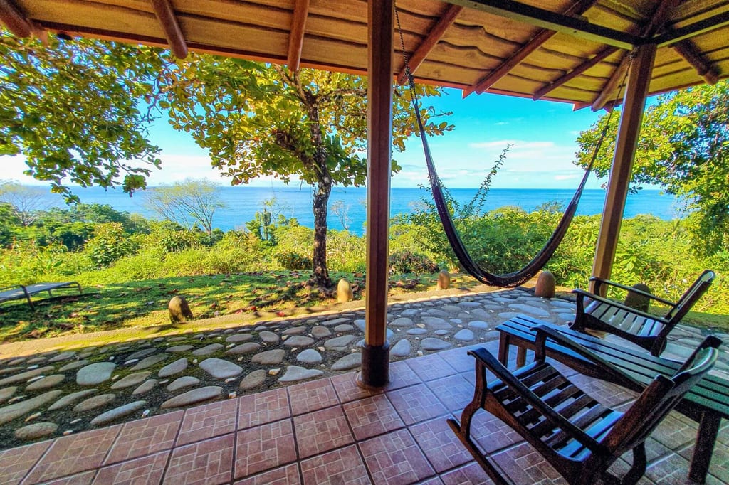 Veranda mit Hängematte und Holzstühlen, Blick auf grüne Küste und blaues Meer bei Tageslicht.