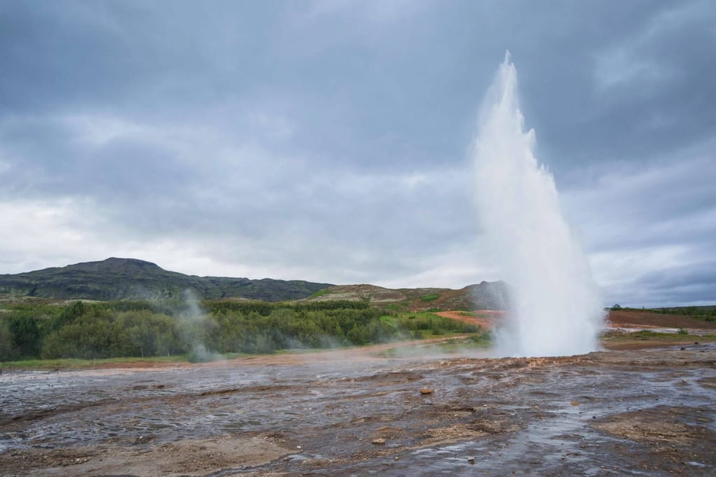 Geysirausbruch mit hoher Wasserfontäne und dampfender, feuchter Landschaft vor bewölkten Hügeln.