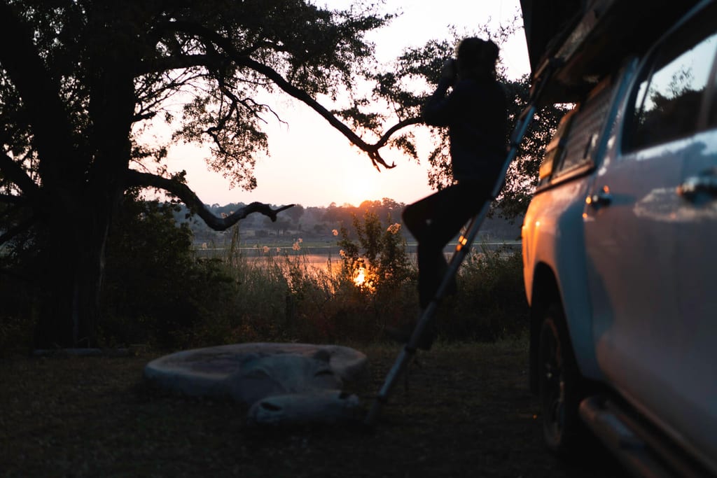 Silhouette einer Person auf einer Leiter neben einem Geländewagen beim Sonnenaufgang über einem See.