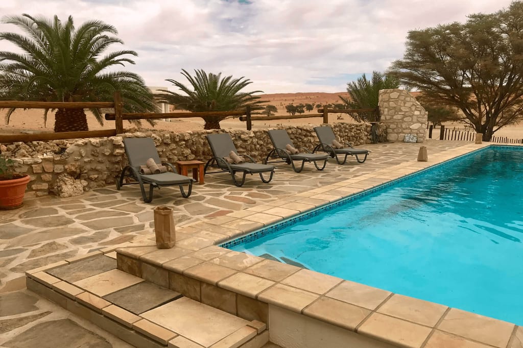 Pool mit Liegestühlen vor Palmen und sandiger Landschaft unter bewölktem Himmel