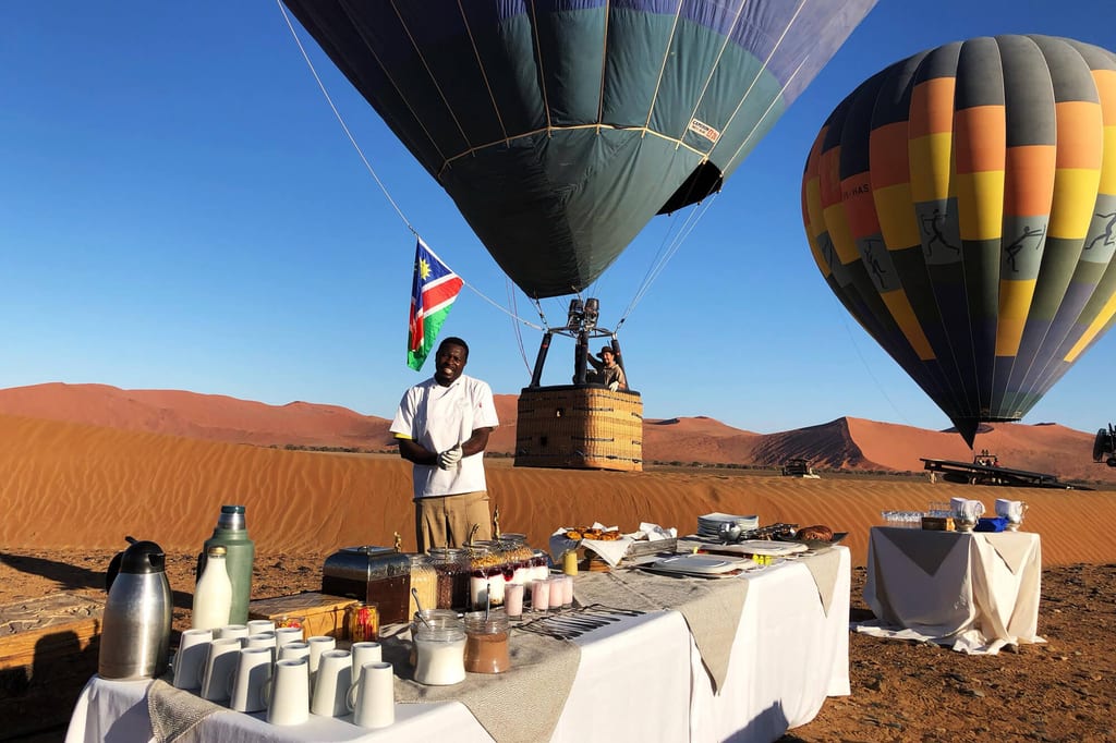 Früher Morgen: Heißluftballons über roten Sanddünen, Frühstücksbuffet und Mitarbeiter im Vordergrund.