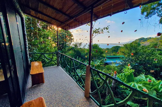 Überdachte Veranda mit Holzbänken und Meerblick über tropische Vegetation im warmen Abendlicht.