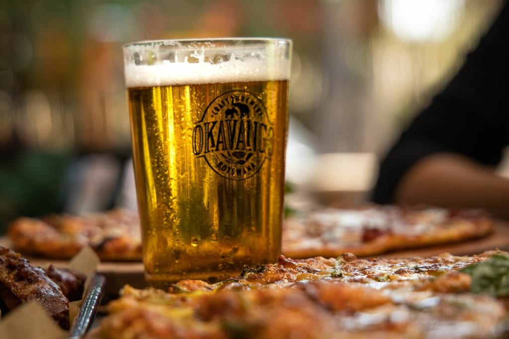 Nahaufnahme eines Bierglases mit Schaumkrone und Pizzen im Vordergrund in gemütlicher Restaurantatmosphäre.