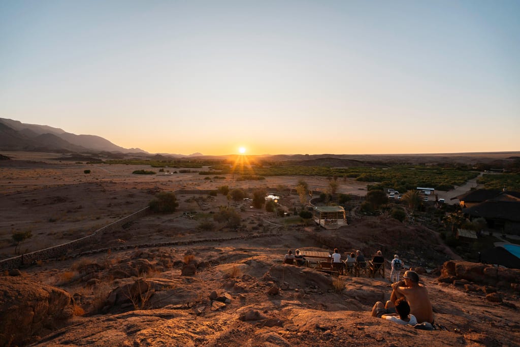 Menschen sitzen auf einem Felsen und schauen im warmen Abendlicht auf eine weite, trockene Ebene mit Sonne am Horizont.