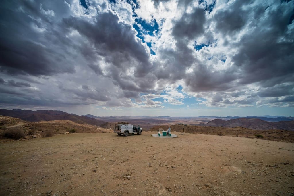Weites trockenes Plateau mit Geländewagen, Person an einer Betonplattform und dramatischen Wolken am Himmel.
