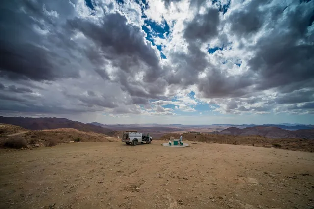 Weites trockenes Plateau mit Geländewagen, Person an einer Betonplattform und dramatischen Wolken am Himmel.