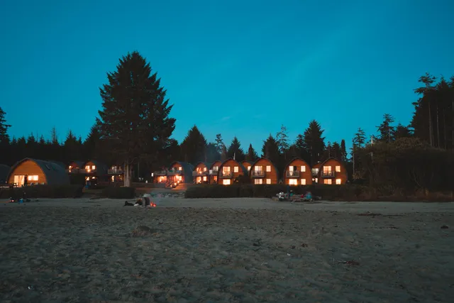 Breiter Sandstrand mit kleinen Lagerfeuern und beleuchteten Holzhütten am Waldrand in der Dämmerung.