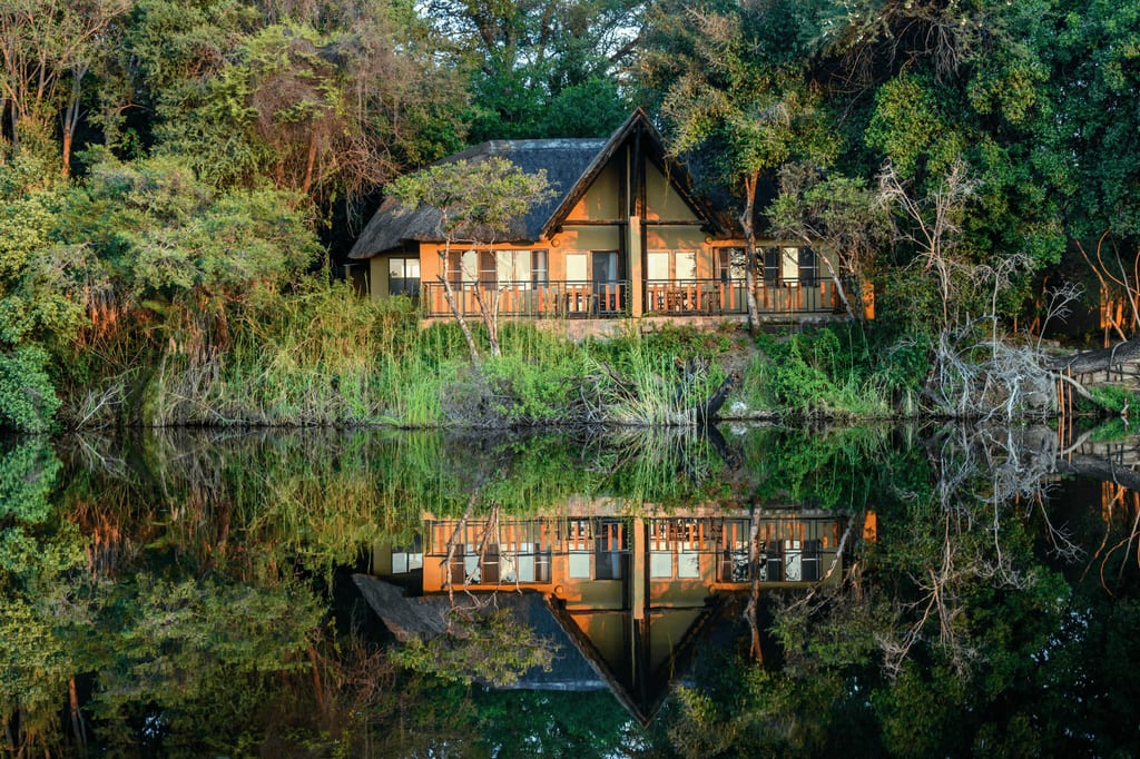 Strohdachhaus mit Veranda spiegelt sich ruhig im Wasser, umgeben von Bäumen und Ufervegetation.