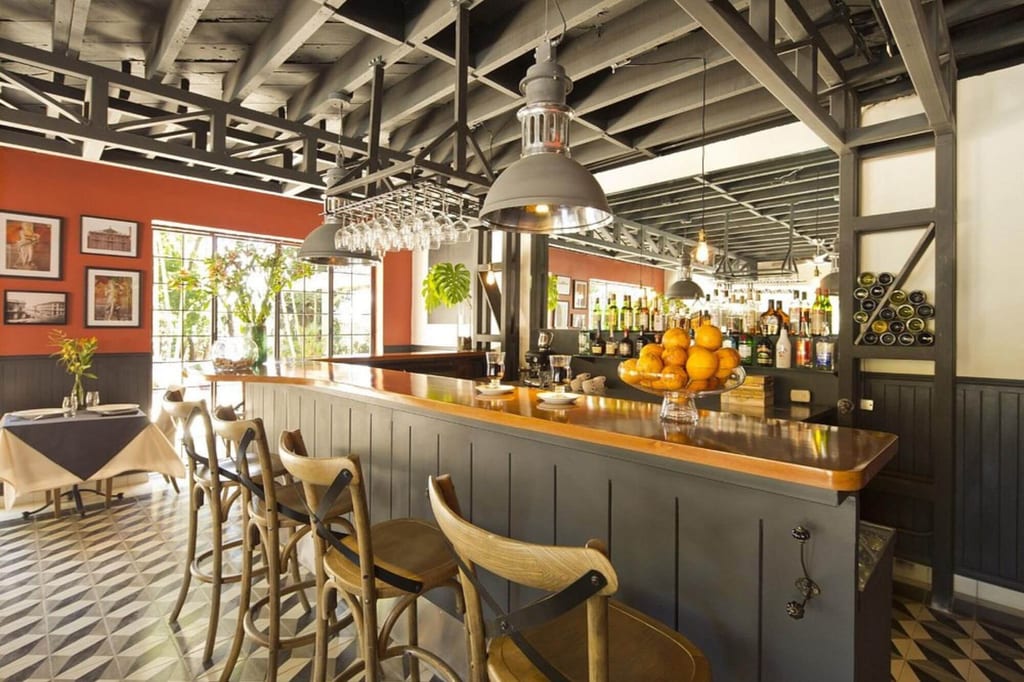 Gemütliche Bar mit langer Holztheke, Hockern und einer Schale Orangen, Tageslicht von großen Fenstern