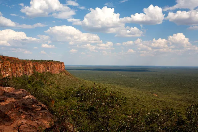 Blick von einem Felsplateau über weite Savannenebene mit roter Klippe und Cumuluswolken