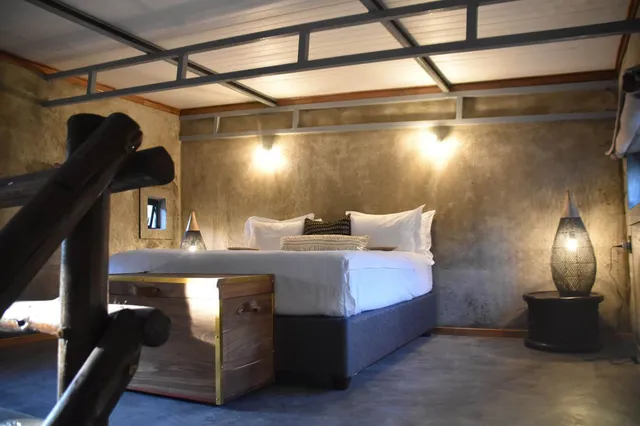 Rustikales Schlafzimmer mit Doppelbett, Holzkoffer und warmen Lampen an Betonwänden.
