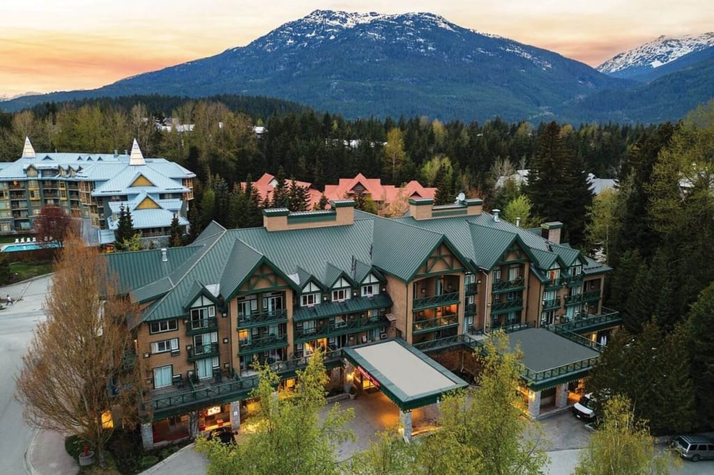 Arealaufnahme eines Hotels mit grünem Dach im Wald vor schneebedeckten Bergen bei Abendlicht.