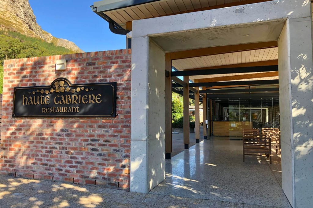 Eingangsbereich des Restaurants mit Schild auf Backsteinwand, überdachter Lobby und Berg im Hintergrund.