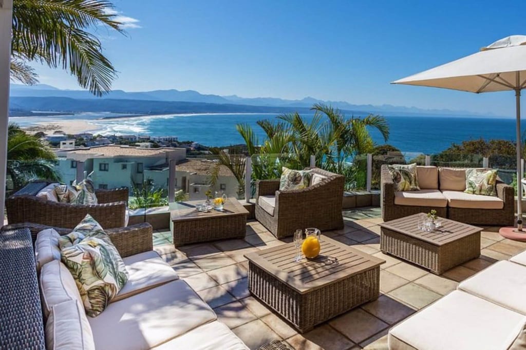 Möblierte Terrasse mit Rattan-Sitzgruppen, Palmen und weitem Meerblick bei klarem Sonnenschein.