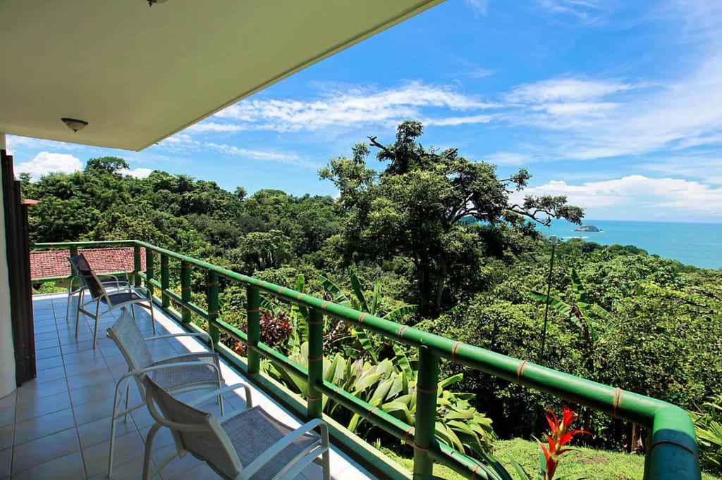 Balkon mit Stühlen und Geländer über tropischem Regenwald mit Blick auf das Meer und blauen Himmel.