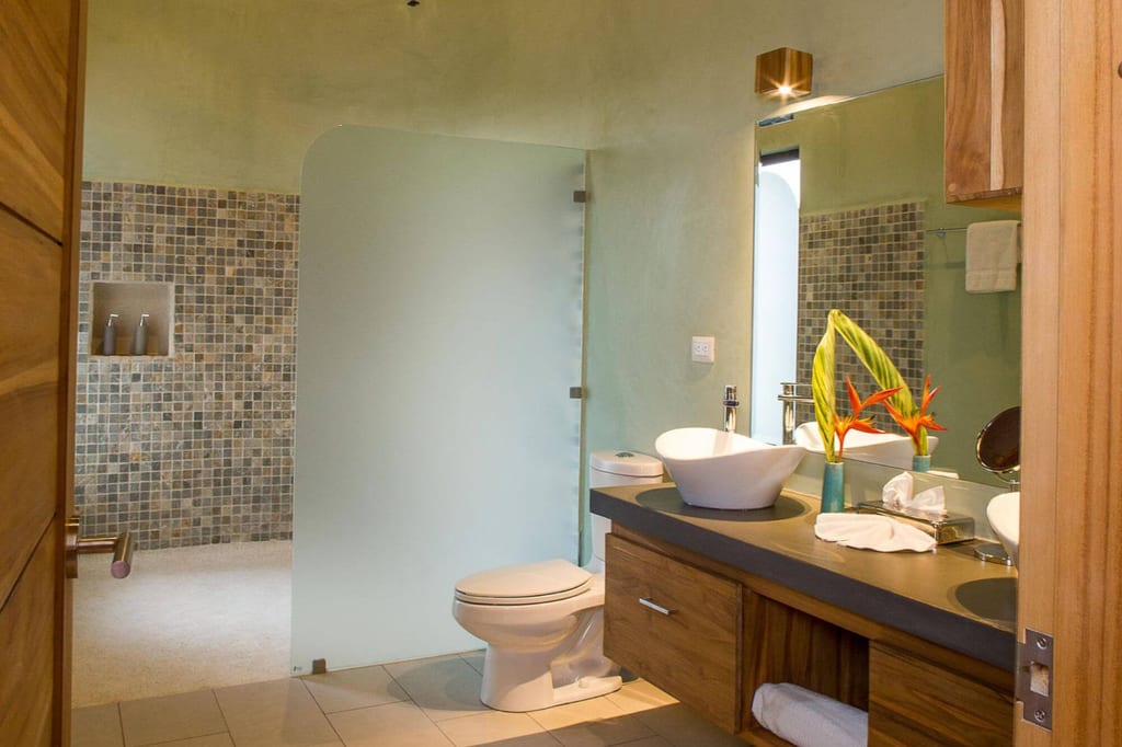 Modernes Badezimmer mit Waschbecken, Toilette, Milchglasscheibe und Mosaikfliesen, Dekoration mit orangefarbenen Blüten.