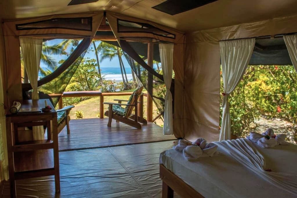 Innenraum eines Strandzelts mit Bett und Veranda, Blick auf Palmen und Meer bei Tageslicht.