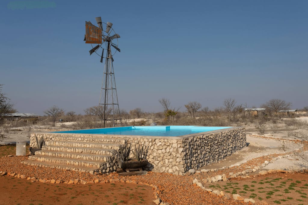 Erhöhtes Schwimmbecken auf Steinpodest mit Windrad in trockener Buschlandschaft bei klarem Tageslicht.