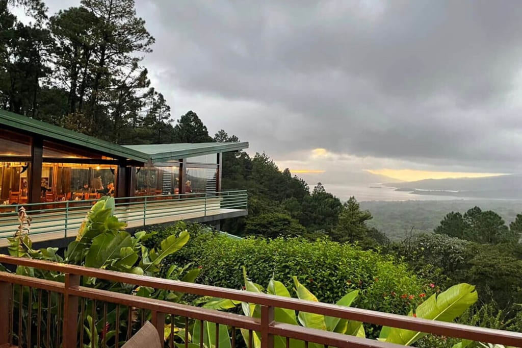 Terrasse mit tropischen Pflanzen vor beleuchteter Lodge und dunstiger Küstenlandschaft unter Wolken