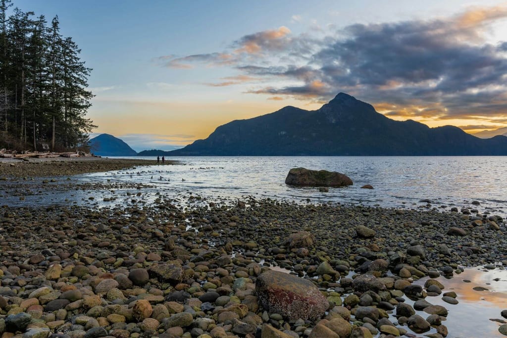 Kieseliger Uferstrand mit Felsen, Nadelwald links und Bergen im warmen Licht der Abenddämmerung