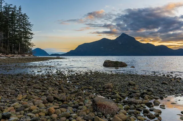 Kieseliger Uferstrand mit Felsen, Nadelwald links und Bergen im warmen Licht der Abenddämmerung