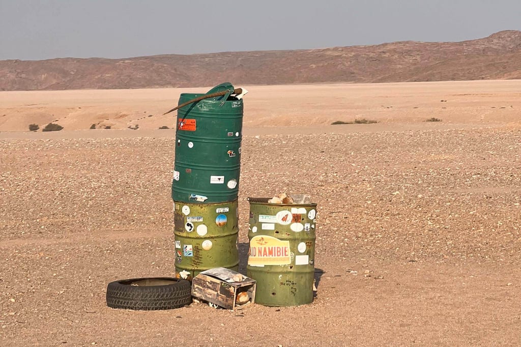Drei bemalte Metallfässer mit Reifen und Kiste in einer kargen Wüstenlandschaft bei hellem Nachmittagslicht.