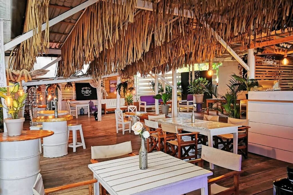 Gemütliche Strandbar mit Strohdach, Holz‑tischen und warmem Licht, Innenperspektive vom vorderen Tisch aus.