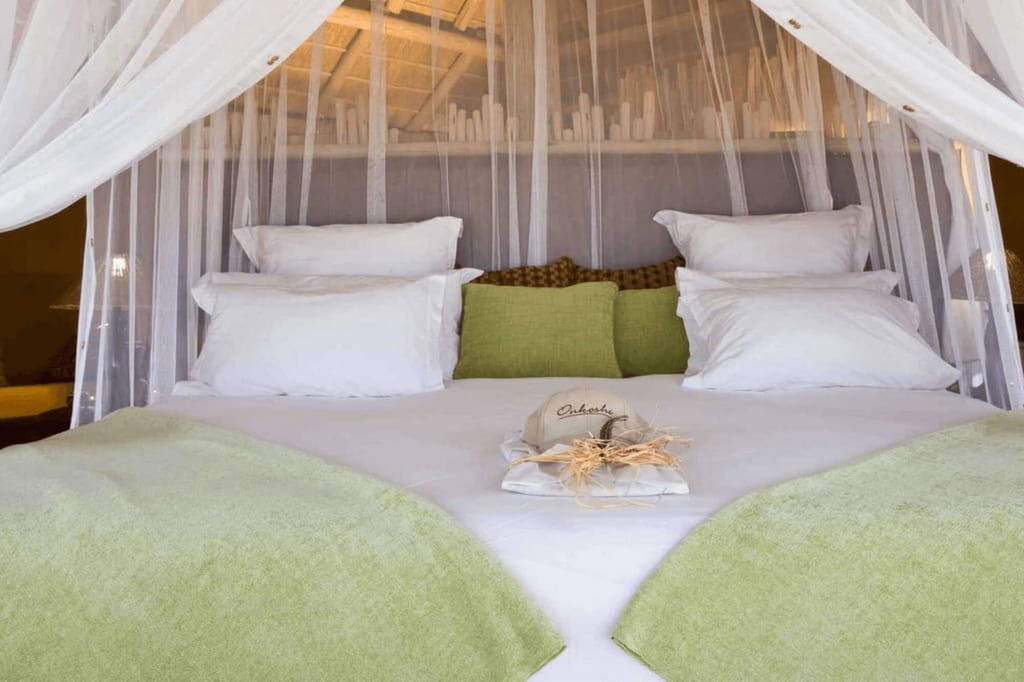 Gemachtes Bett mit weißem Moskitonetz, grünen Überwürfen und dekorativem Hut in der Mitte.