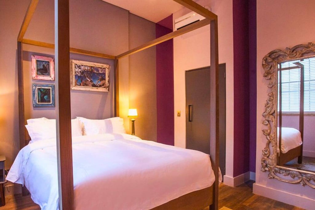 Gemütliches Schlafzimmer mit vierpfostenbett, weißer Bettwäsche, lila Wänden und großem verzierten Spiegel.