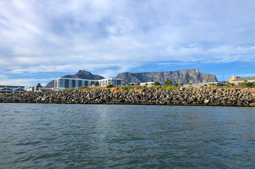 Blick vom Wasser auf felsiges Ufer, Gebäude und markante Bergformation unter wolkigem Himmel.