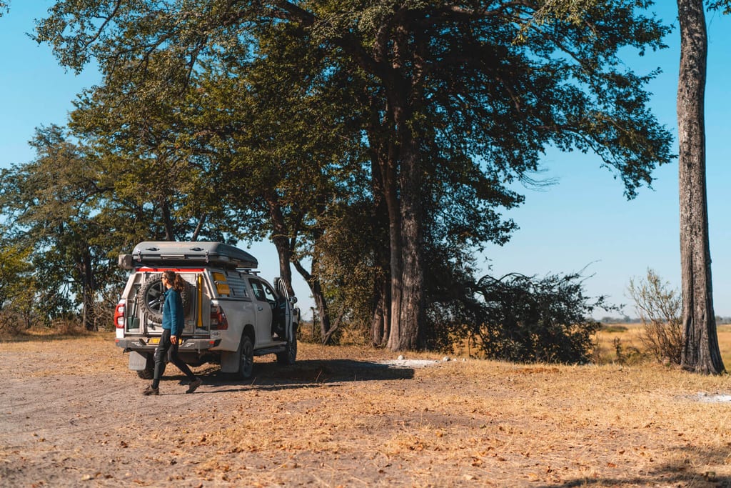 Geländewagen steht unter großen Bäumen auf trockener Graslandschaft, Person geht vom Heck weg.