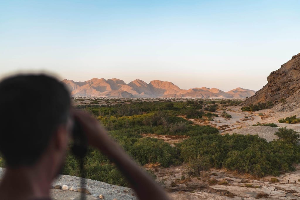 Weite Flusslandschaft mit Büschen und Bergen im warmen Abendlicht, unscharfe Person mit Fernglas im Vordergrund.