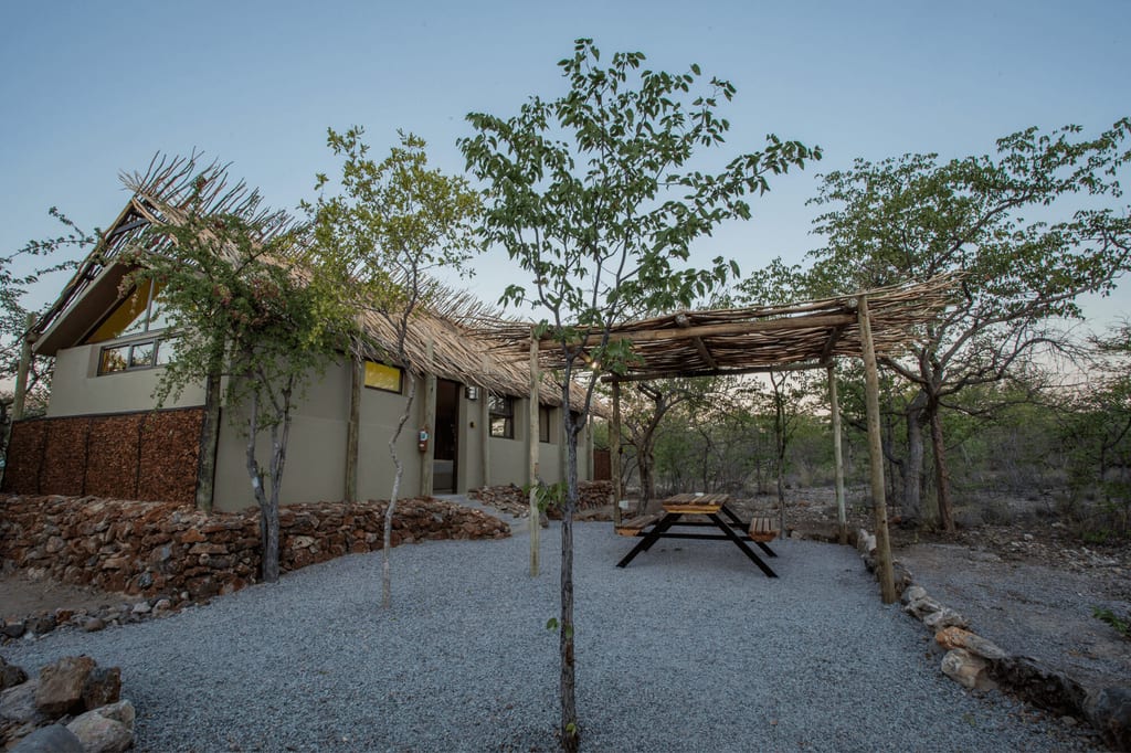 Lodge mit strohgedecktem Dach, Pergola und Picknickbank in trockener Buschlandschaft bei Abendlicht.