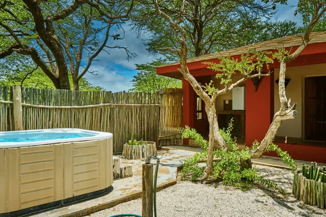 Innenhof einer Lodge mit Whirlpool, roten Wänden, Holzzaun und knorrigen Bäumen bei Sonnenschein.