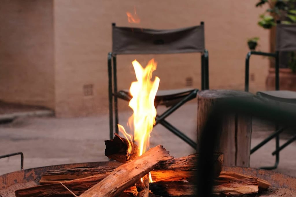 Brennendes Lagerfeuer in einer Feuerstelle vor Campingstühlen und Baumstumpf im warmen Abendlicht.