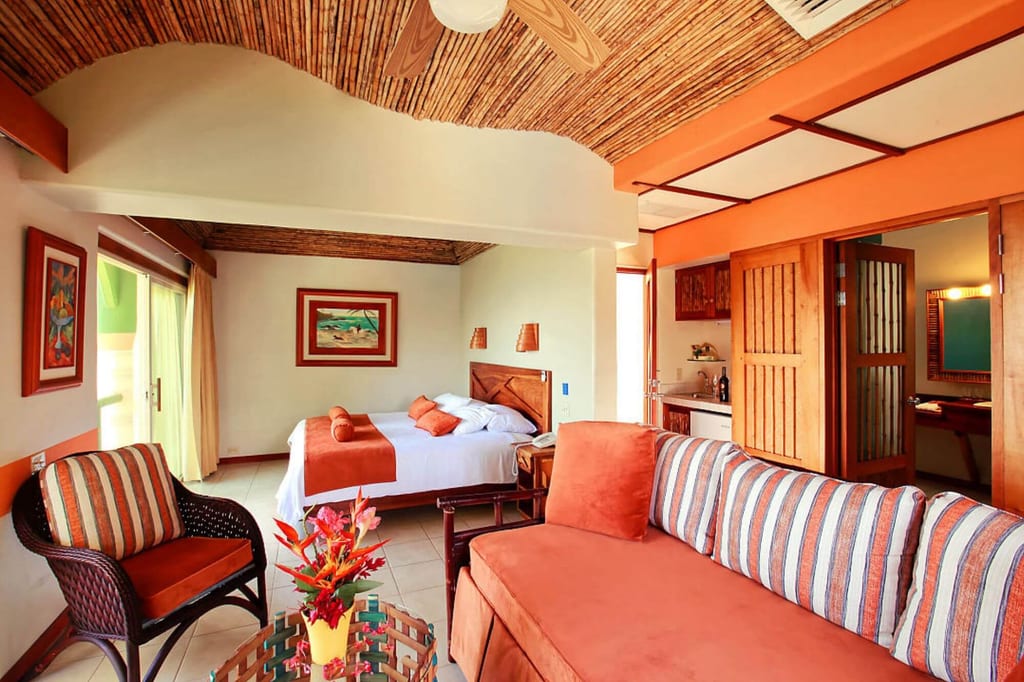 Helles Hotelzimmer mit Wohnbereich, Bett, Sofa und Terrassentür in warmen Orangetönen.