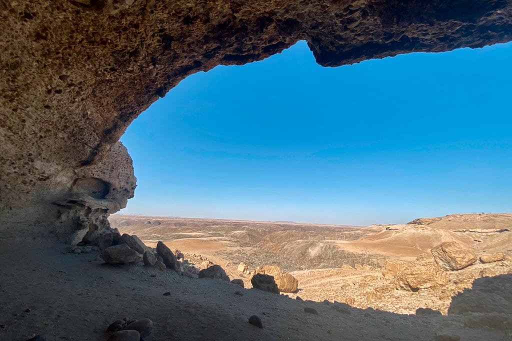 Blick aus einer Felsgrotte auf weite, trockene Wüstenlandschaft unter klarem blauen Himmel.