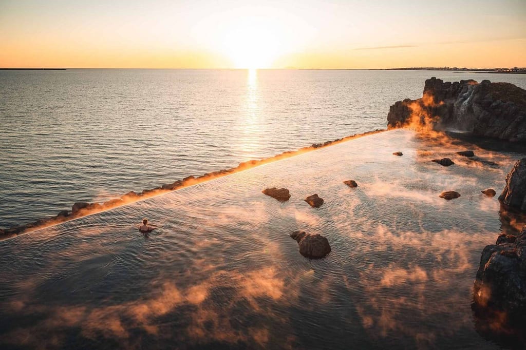 Erhöhter Blick auf ein dampfendes Felsbecken am Meer mit einer Person im Wasser bei Sonnenuntergang