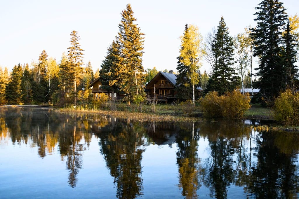 Holzhäuser und Bäume spiegeln sich im ruhigen See bei goldenem Spätnachmittagslicht.