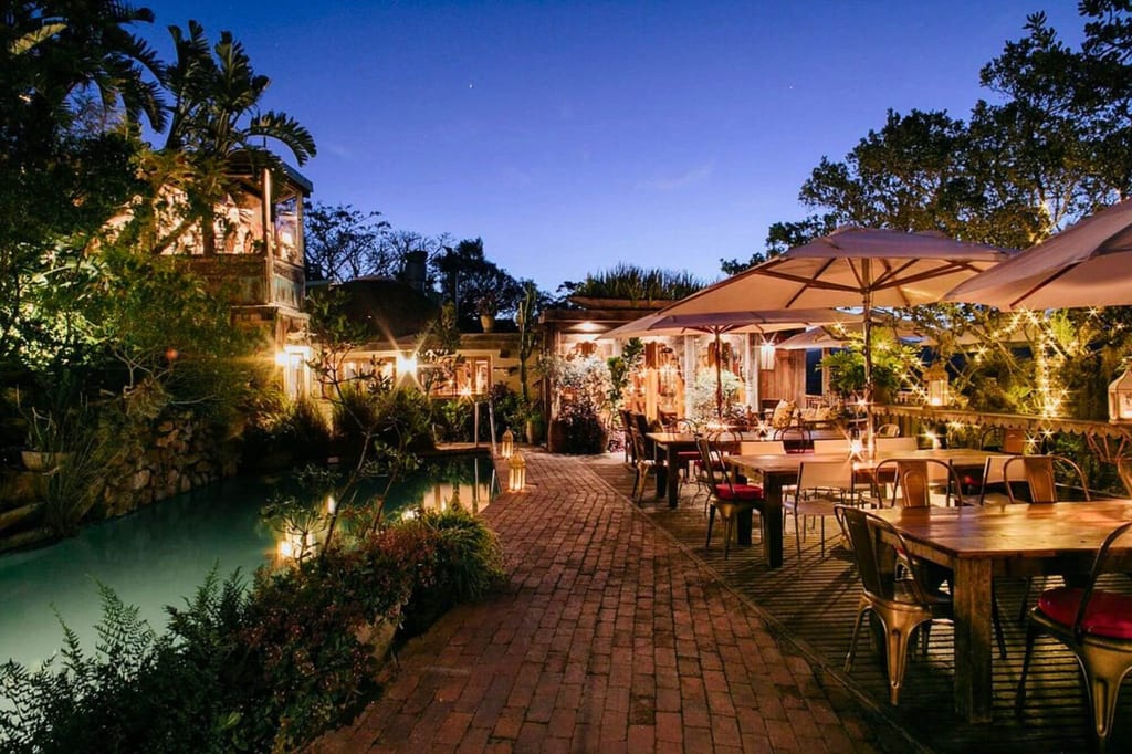 Stimmungsvolle, beleuchtete Außen-Terrasse mit Tischen und Sonnenschirmen neben einem Teich in der Abenddämmerung.