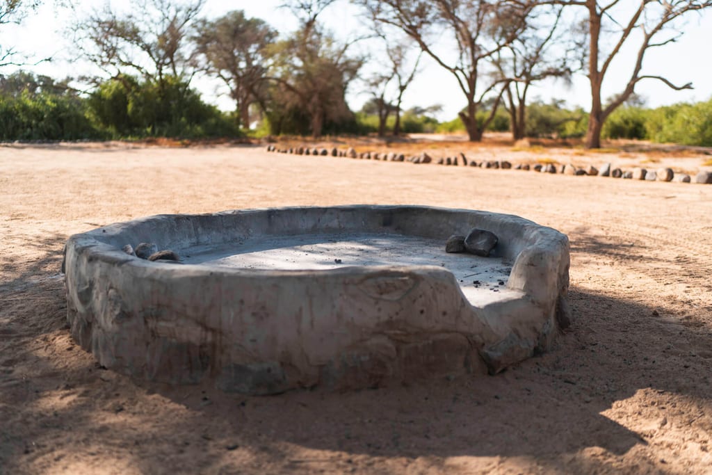 Runde Betonwasserstelle in sandiger, sonniger Landschaft mit Bäumen im Hintergrund.