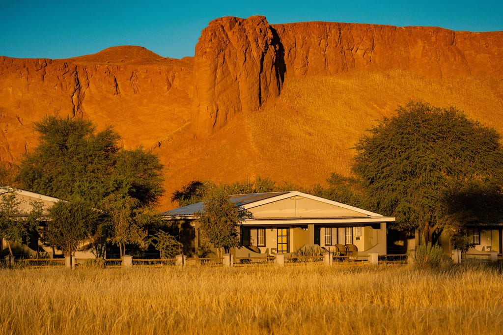 Lodge vor roter Felswand im warmen Licht der Abendsonne, Hochgras und Bäume im Vordergrund.