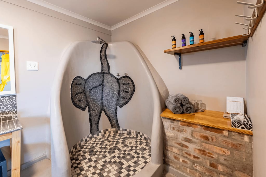 Badezimmer mit runder Dusche und Elefanten-Mosaik, Handtücher und Regal bei warmem Licht.
