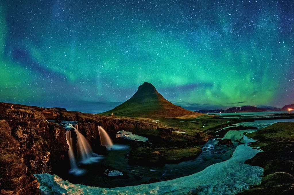 Nächtliche Landschaft mit Kegelfelsen, Wasserfall und grünen Nordlichtern am klaren Sternenhimmel.