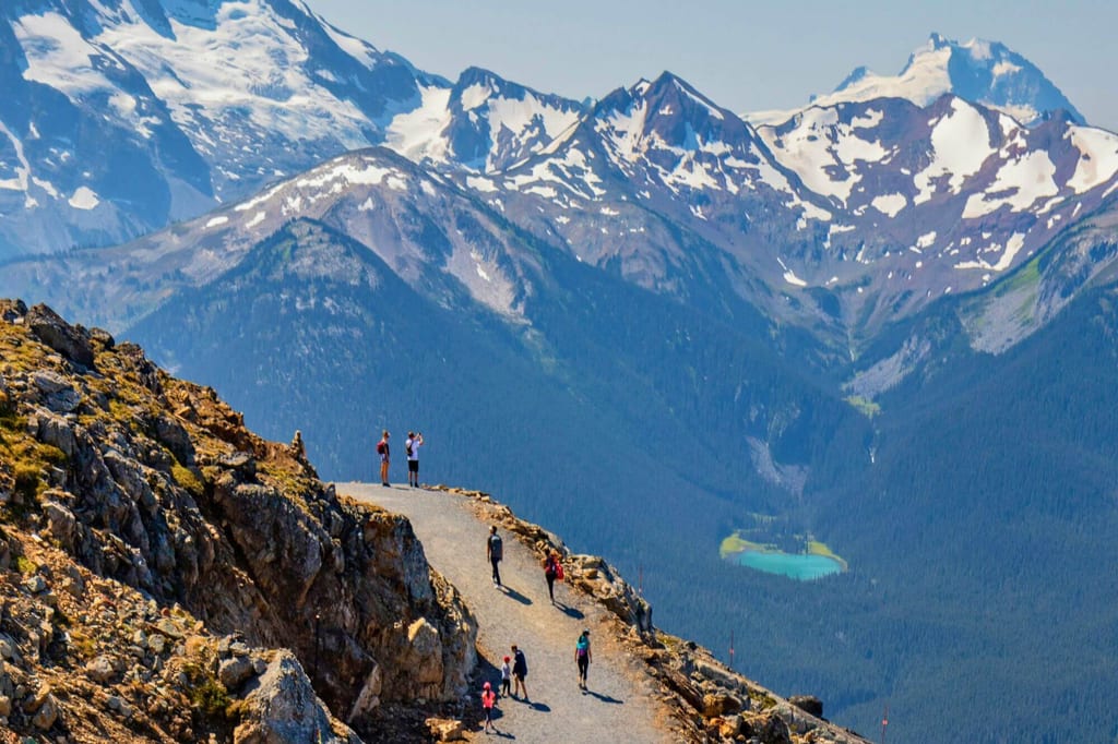 Wanderer auf Aussichtspfad bei Whistler vor schneebedeckten Gipfeln und türkisfarbenem Bergsee