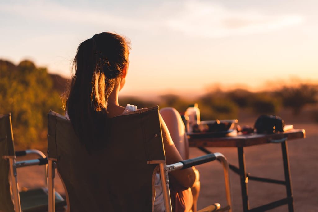 Person von hinten im Campingstuhl bei Sonnenuntergang vor Campingtisch mit Geschirr
