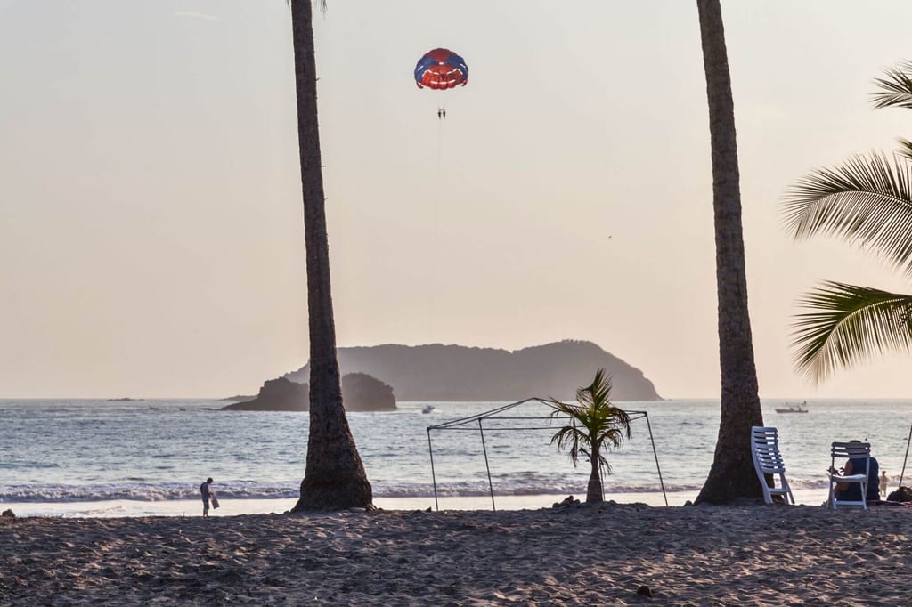 Strand bei Abendlicht mit Palmen, Liegestühlen und einem Parasail über dem Meer