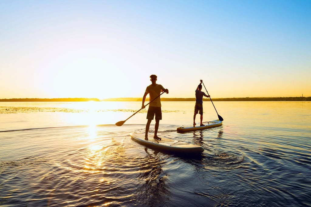 Zwei Personen paddeln auf Stand-up-Paddleboards bei Sonnenuntergang auf einem ruhigen See.