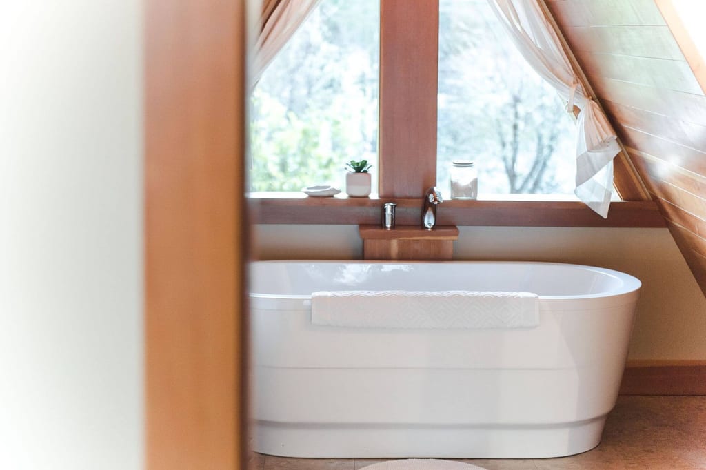 Freistehende weiße Badewanne vor Fenster in gemütlichem Dachzimmer, weiches Tageslicht
