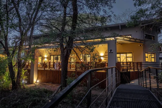 Beleuchtete Holzterrasse eines Hauses bei Abenddämmerung, Vordergrund erhöhter Metallsteg durch Bäume.